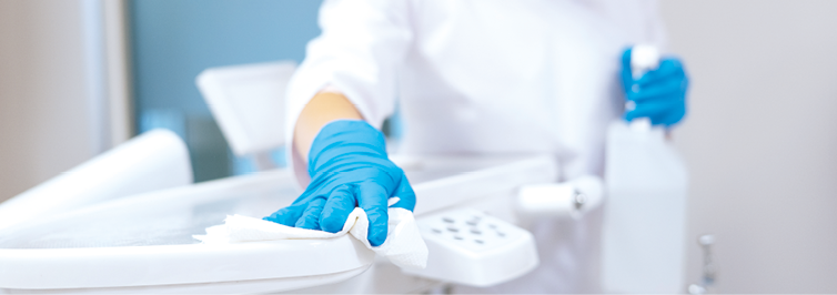 Nurse in protective gloves cleaning work surface at stomatology clinic, sanitizing table with disinfectant spray bottle, washing dental chair with towel, selective focus
